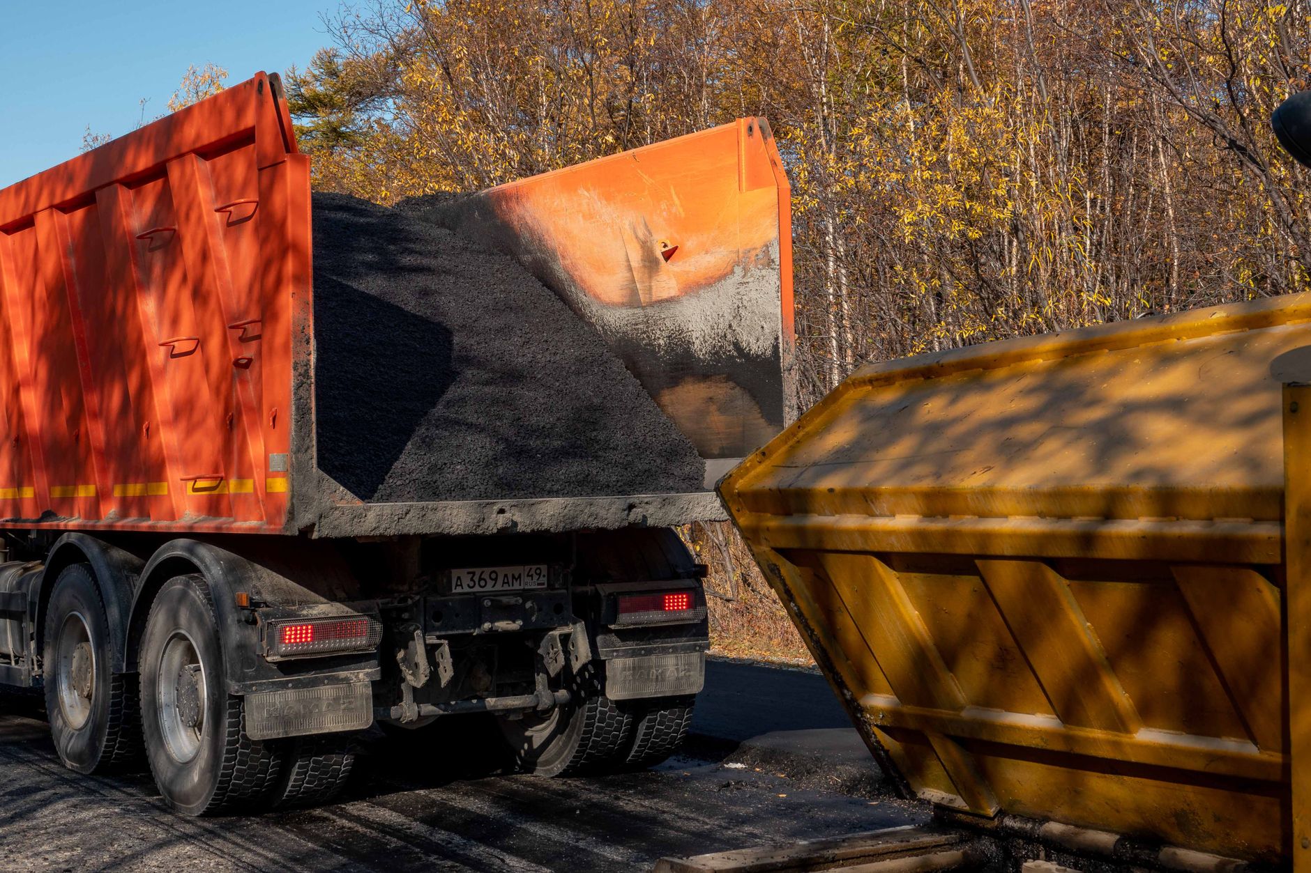 A dump truck unloading asphalt into a construction bin on a sunny autumn day.