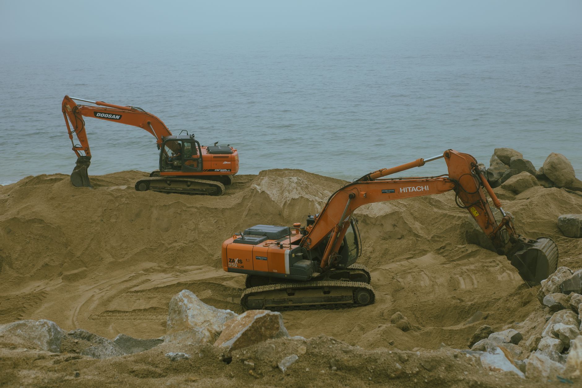 Two excavators operate at a sandy construction site by the sea, moving sand and rock.